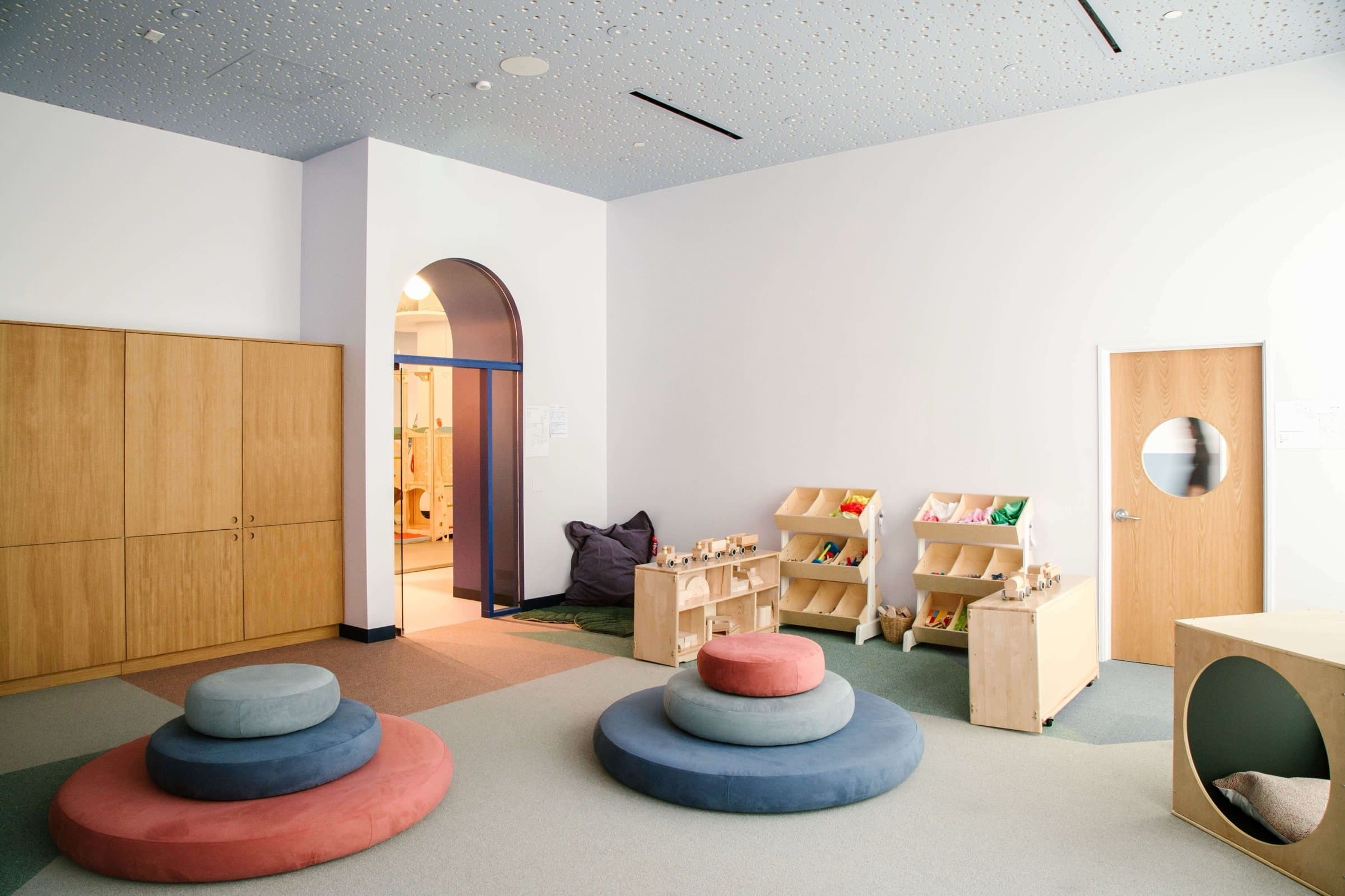 Brella playroom — stacked round floor cushions in coral and blue, white-oak storage, an arched doorway, and a starry acoustic ceiling